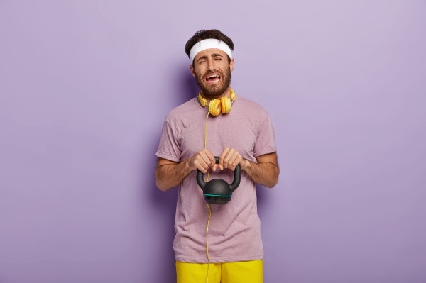 Frustrated lifter struggling with a small kettlebell in front of a purple wall
