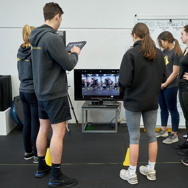 Coach reviews video analysis on a tablet beside athletes in a gym